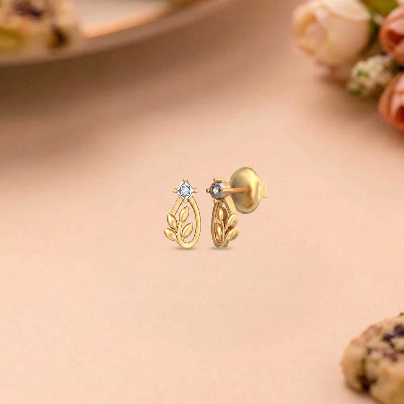 A pair of gold leaf stud earrings, one facing forward with a small light blue flower.