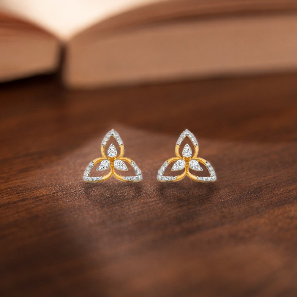 A pair of gold and white gold triangular stud earrings, resting on a dark wooden surface with a blurred open book in the background.