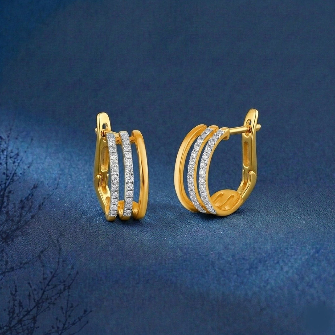A pair of gold hoop earrings with three parallel rows of small clear stones on the front, displayed on a textured dark blue background.