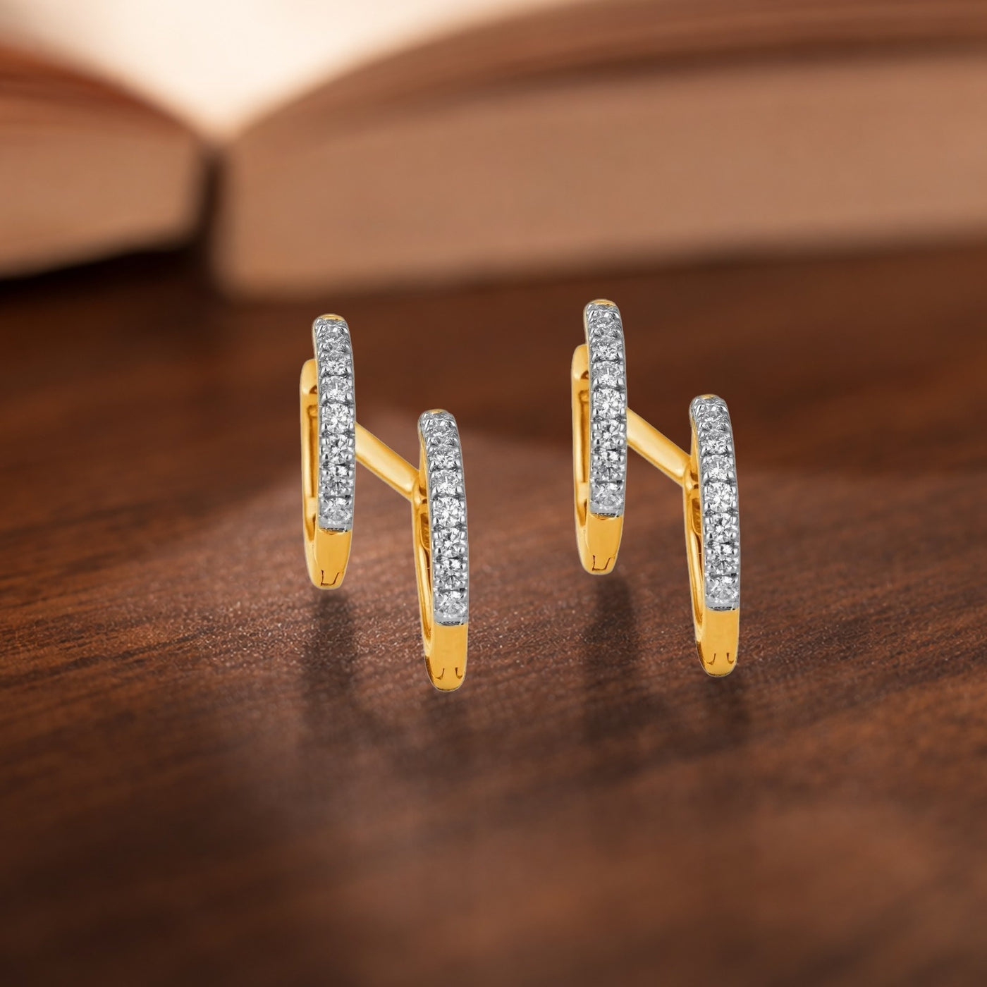 A pair of gold-toned geometric earrings, displayed on a dark wooden surface with a blurred open book in the background.