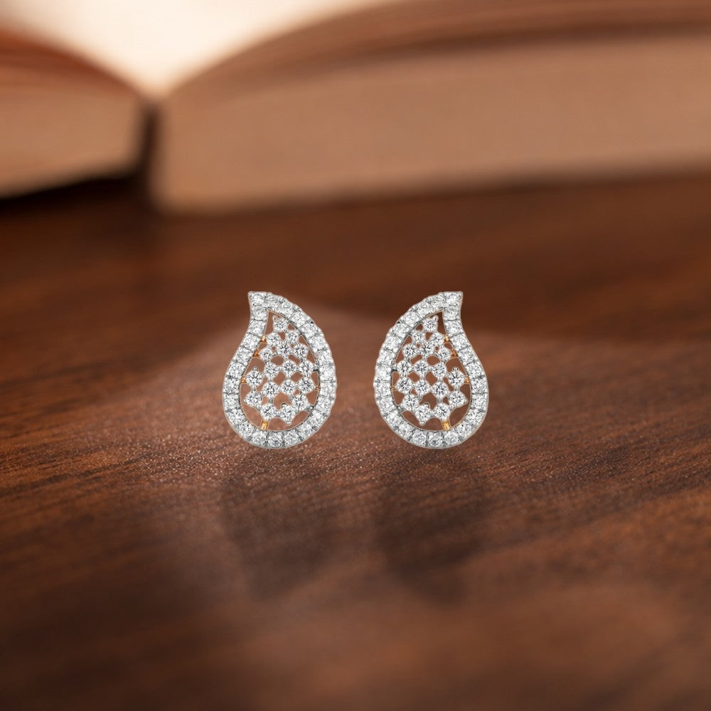 A pair of teardrop-shaped diamond stud earrings paved with clear stones, resting on a dark wooden surface with a blurred open book in the background.