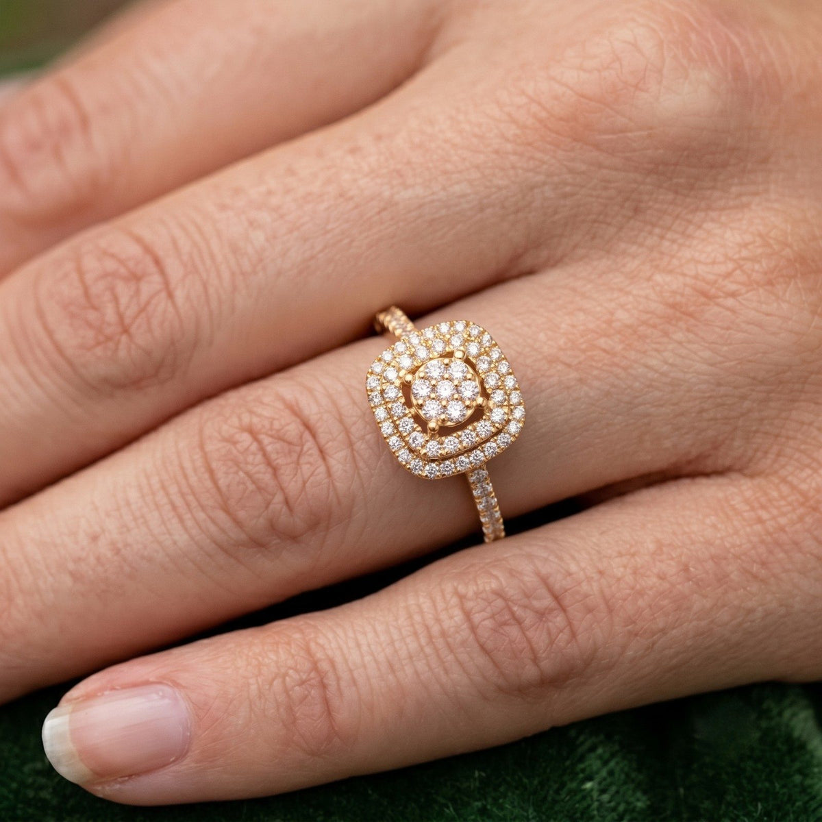 A close-up of a hand wearing a gold ring with a square diamond center, set against a dark green textured background.