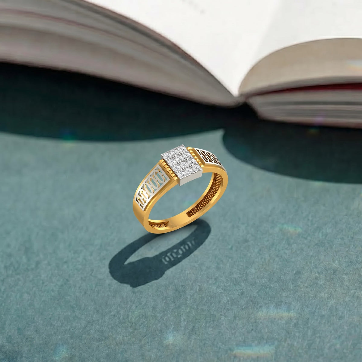 A gold ring with a rectangular white stone cluster, resting on a textured teal surface with an open book blurred in the background.