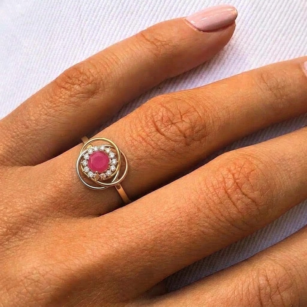 A close-up of a hand wearing a rose gold ring with a central round ruby gemstone surrounded by a halo of small clear stones.