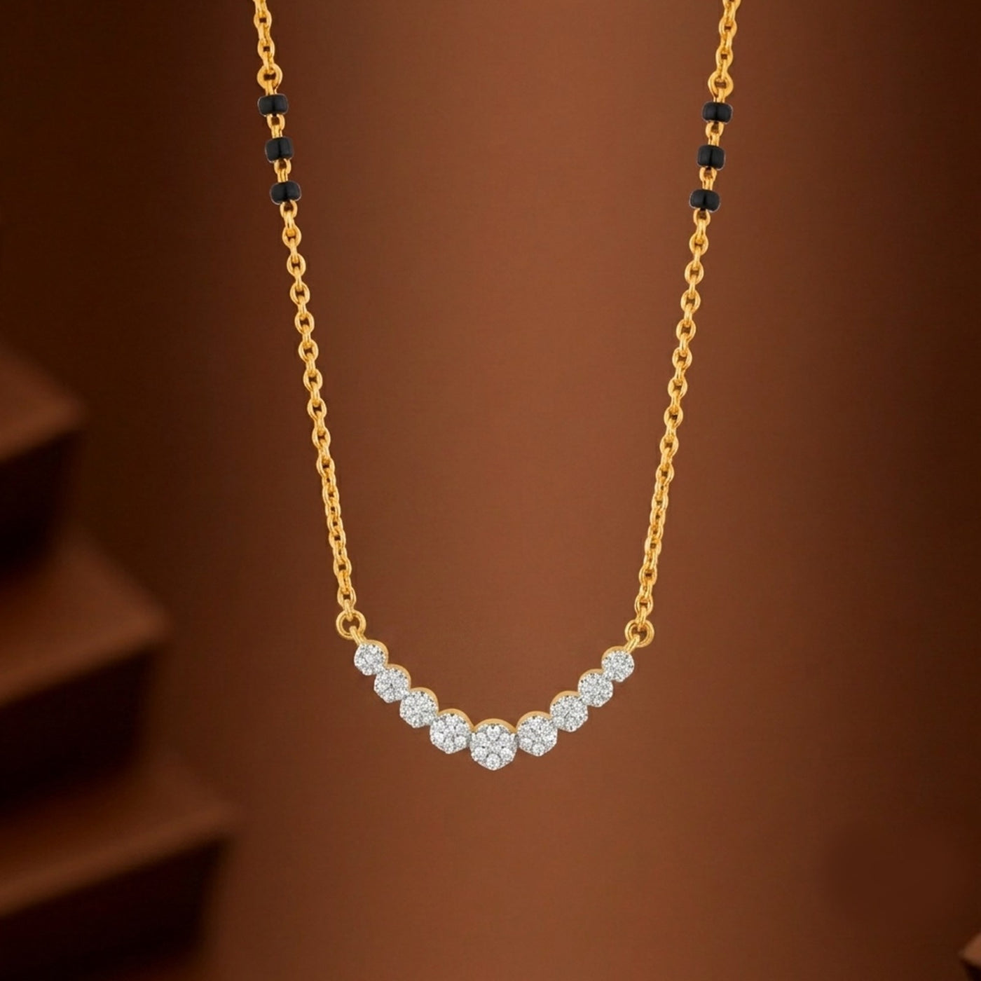 A yellow gold and diamond mangalsutra featuring a curved row of clear round-cut stones at the center, with small black bead accents on the chain near the top, shown against a warm brown background.