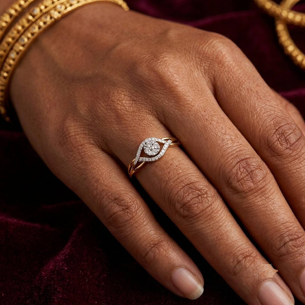 A close-up of a hand wearing a gold ring with a diamond eye motif, set against a dark burgundy velvet background.
