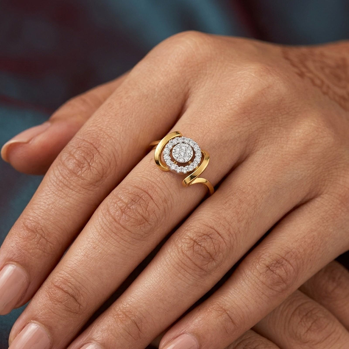 A close-up of a hand wearing a gold ring with a circular diamond cluster, set against a softly blurred dark blue background.