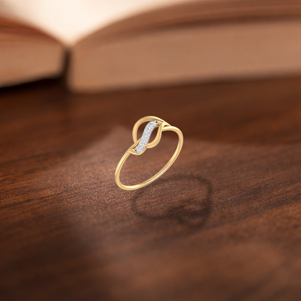 A gold ring with a curved, intertwined design, resting on a dark wooden surface with a blurred book in the background.