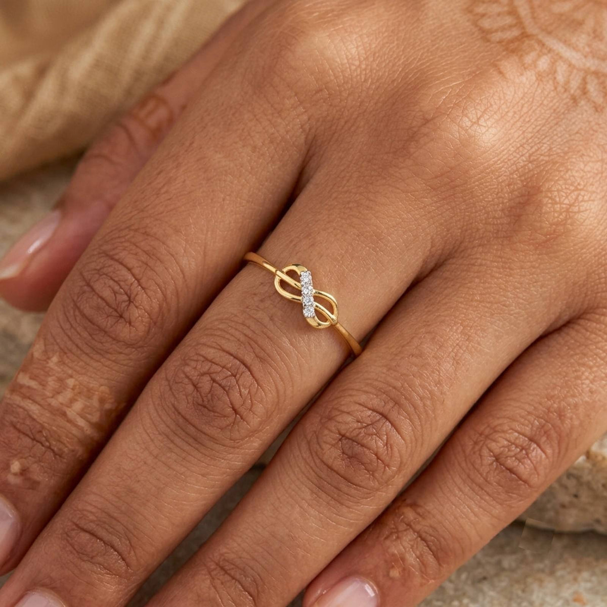 A gold infinity ring with a small clear stone on the center of the band, shown worn on a person's finger.