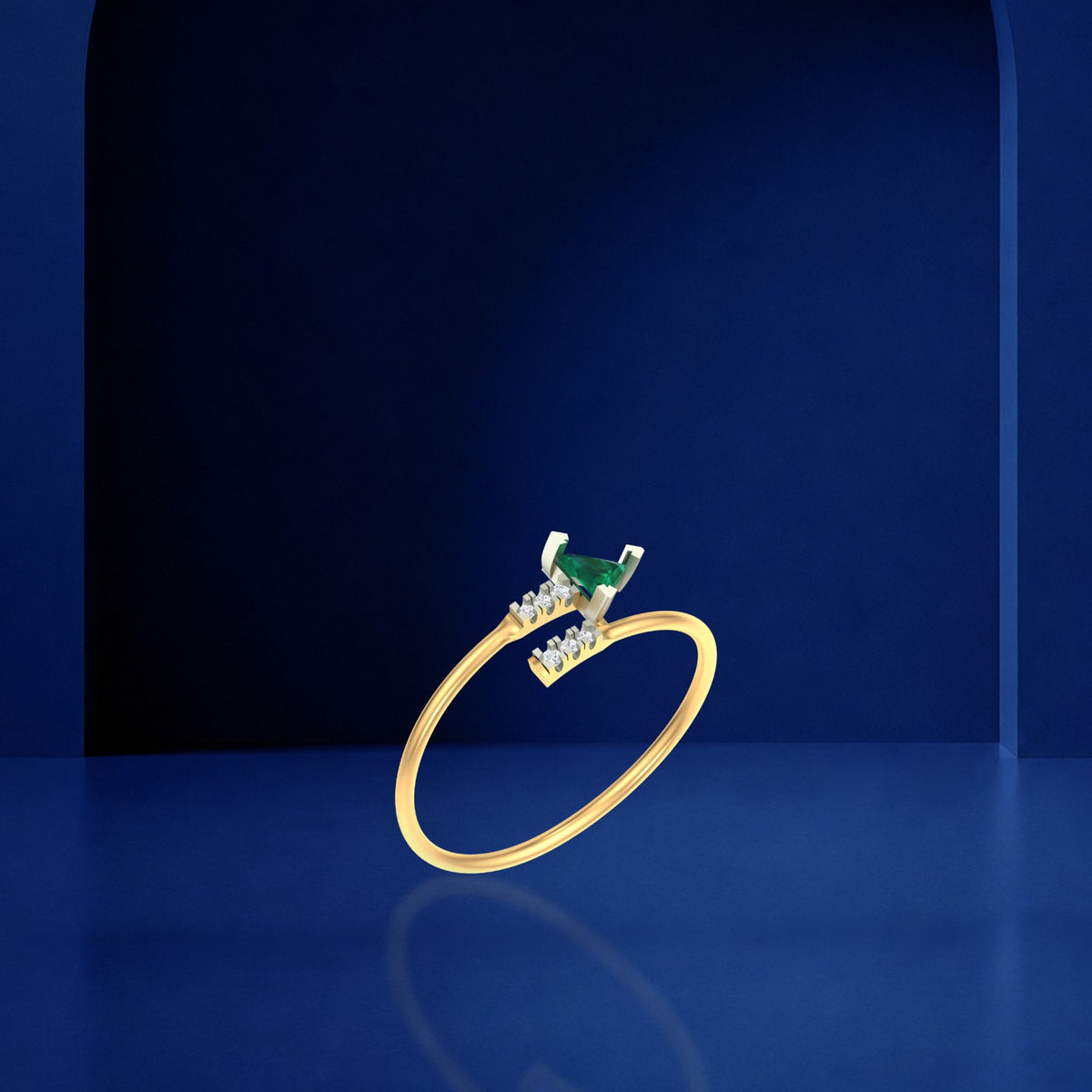 A gold ring with a small emerald triangle-shaped gemstone, displayed on a dark blue reflective surface against a matching blue background.