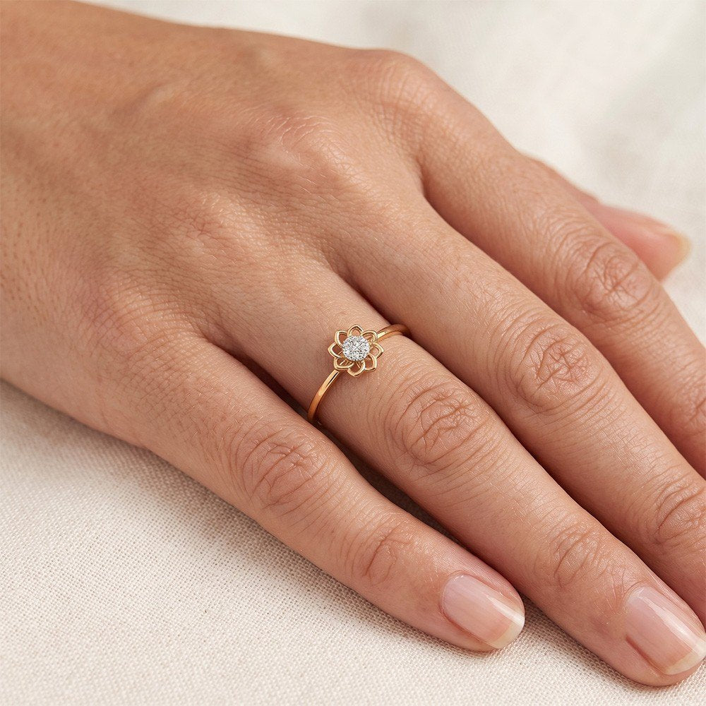 A delicate gold ring with a flower-shaped setting, shown worn on a person's finger against a soft beige fabric background.
