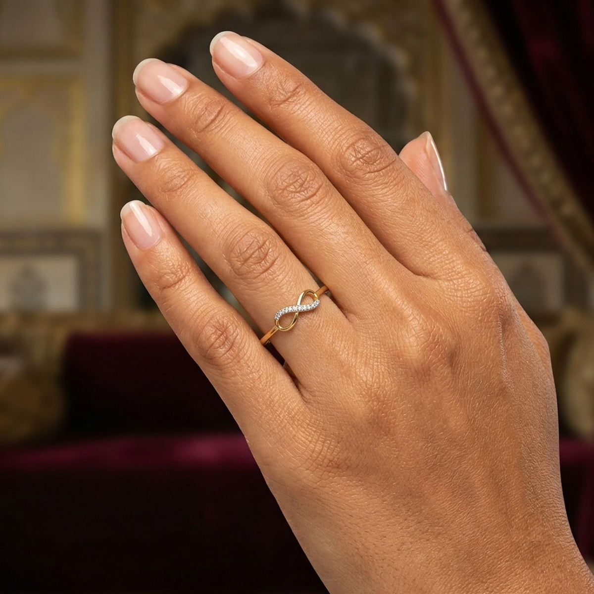 A close-up of a hand wearing a delicate infinity-style ring, set against an elegant, softly blurred interior background.