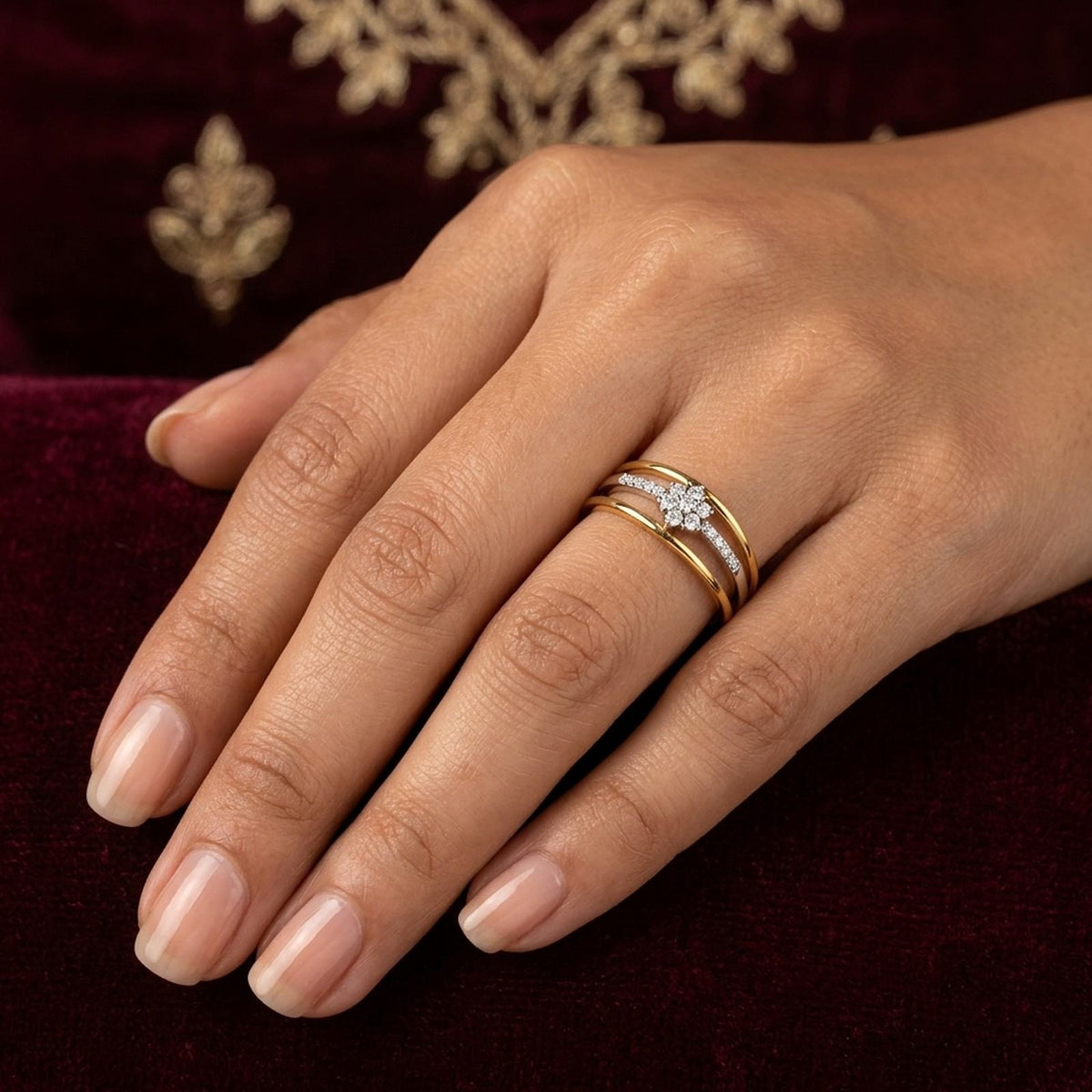 A close-up of a hand wearing a gold ring with a diamond floral center, set against a dark burgundy background.