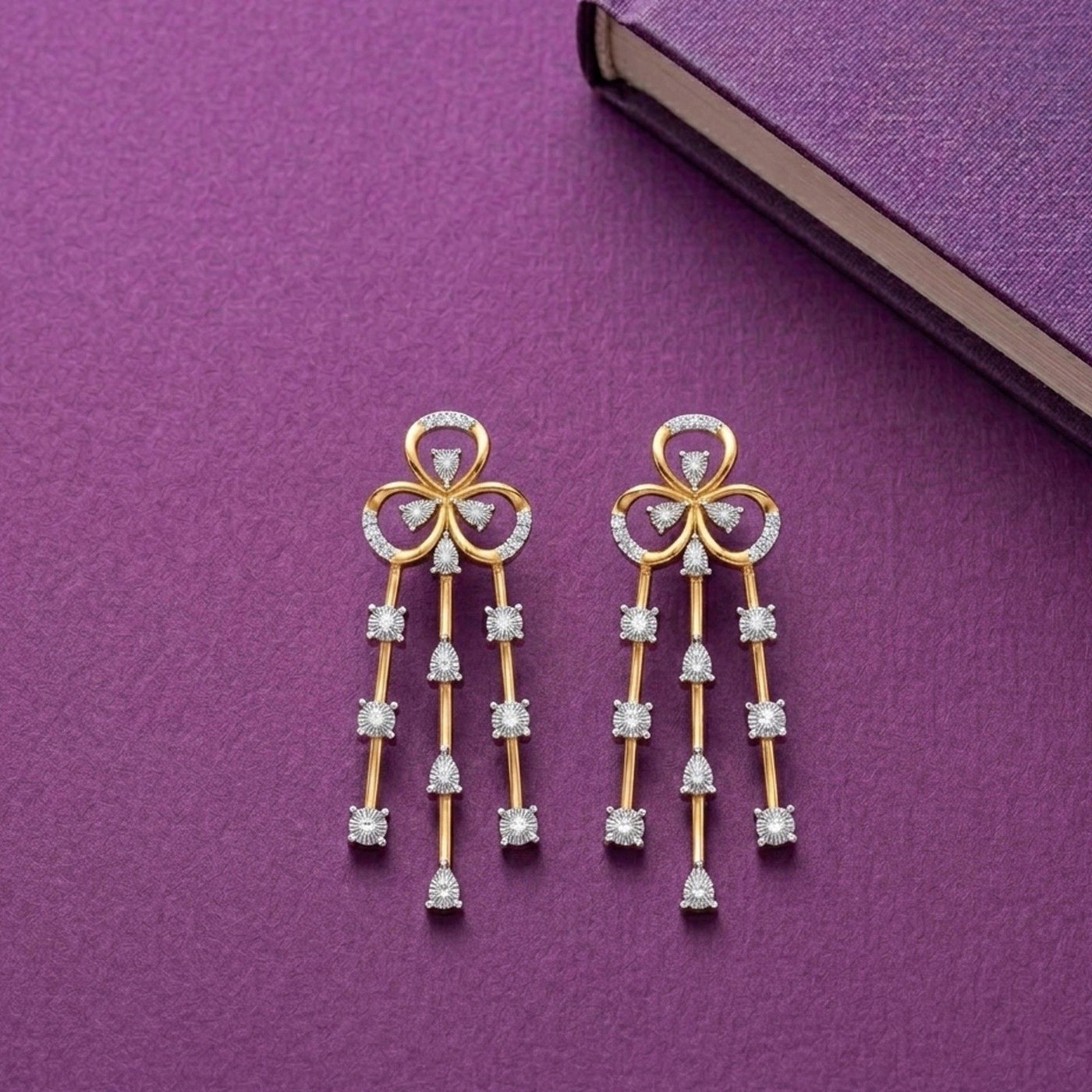 A pair of gold and diamond drop earrings featuring a floral bow design on a purple background