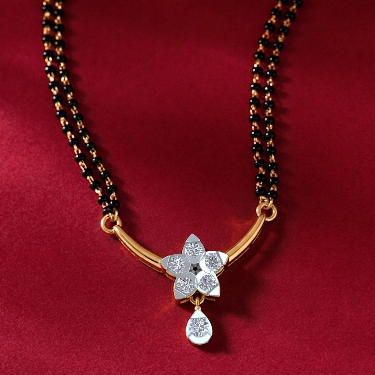 A double-strand black bead mangalsutra with a curved gold featuring a diamond flower and teardrop dangle, resting on dark red fabric