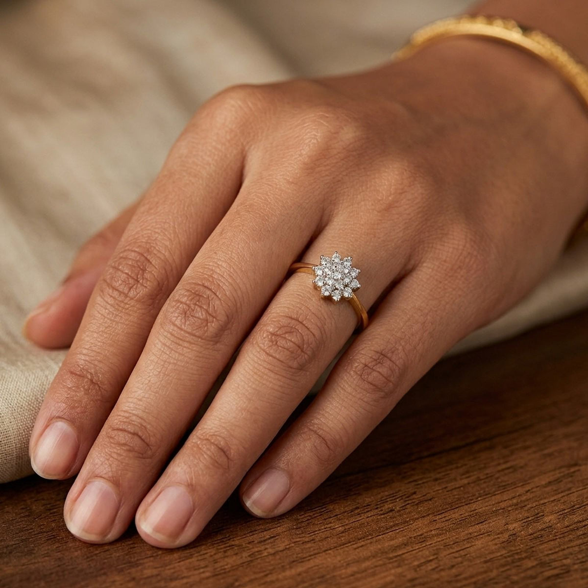 A close-up of a hand wearing a gold ring with a round cluster, resting on a wooden surface with a beige fabric background.