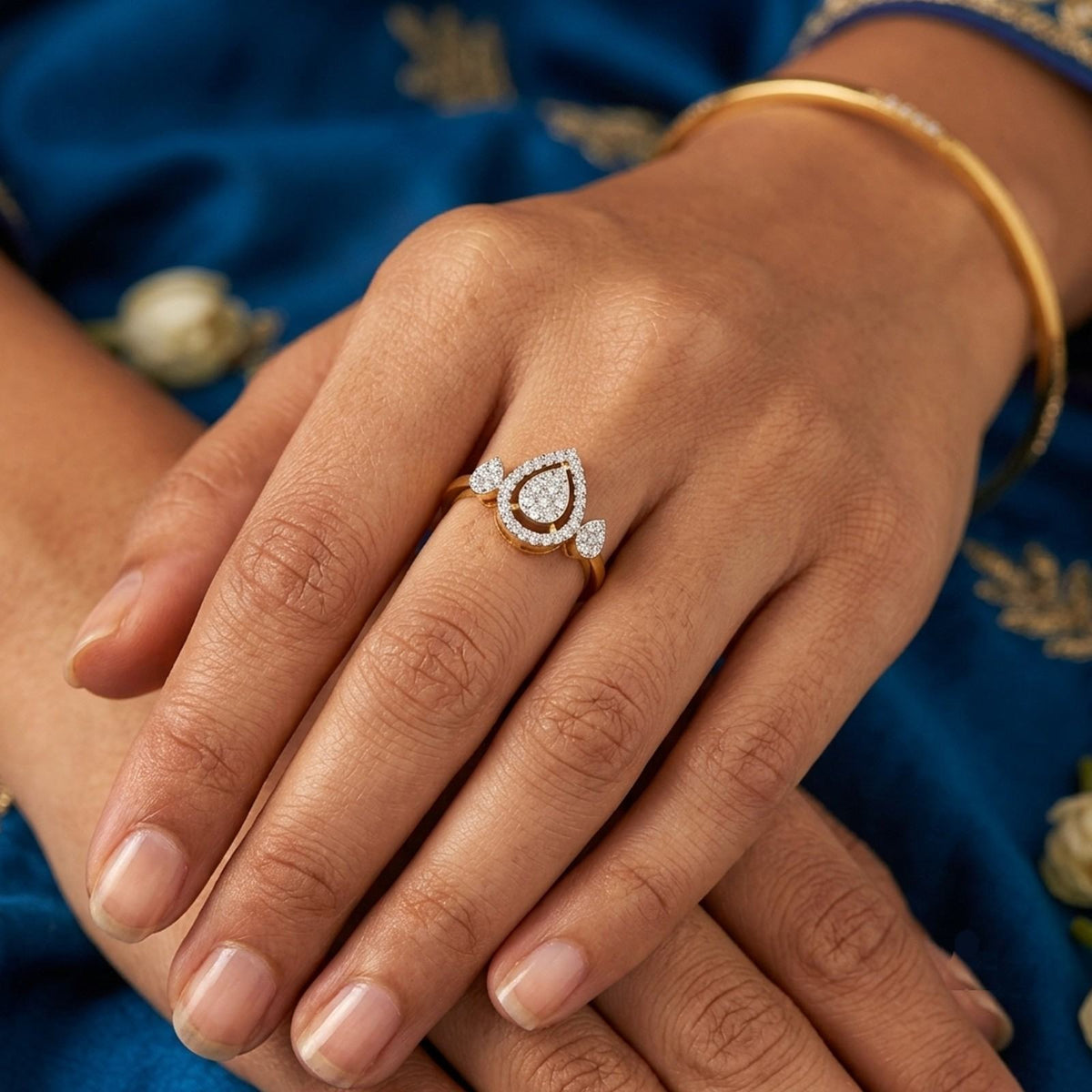 A gold ring with a teardrop-shaped diamond cluster worn on a woman's hand, set against a blue embroidered fabric background.