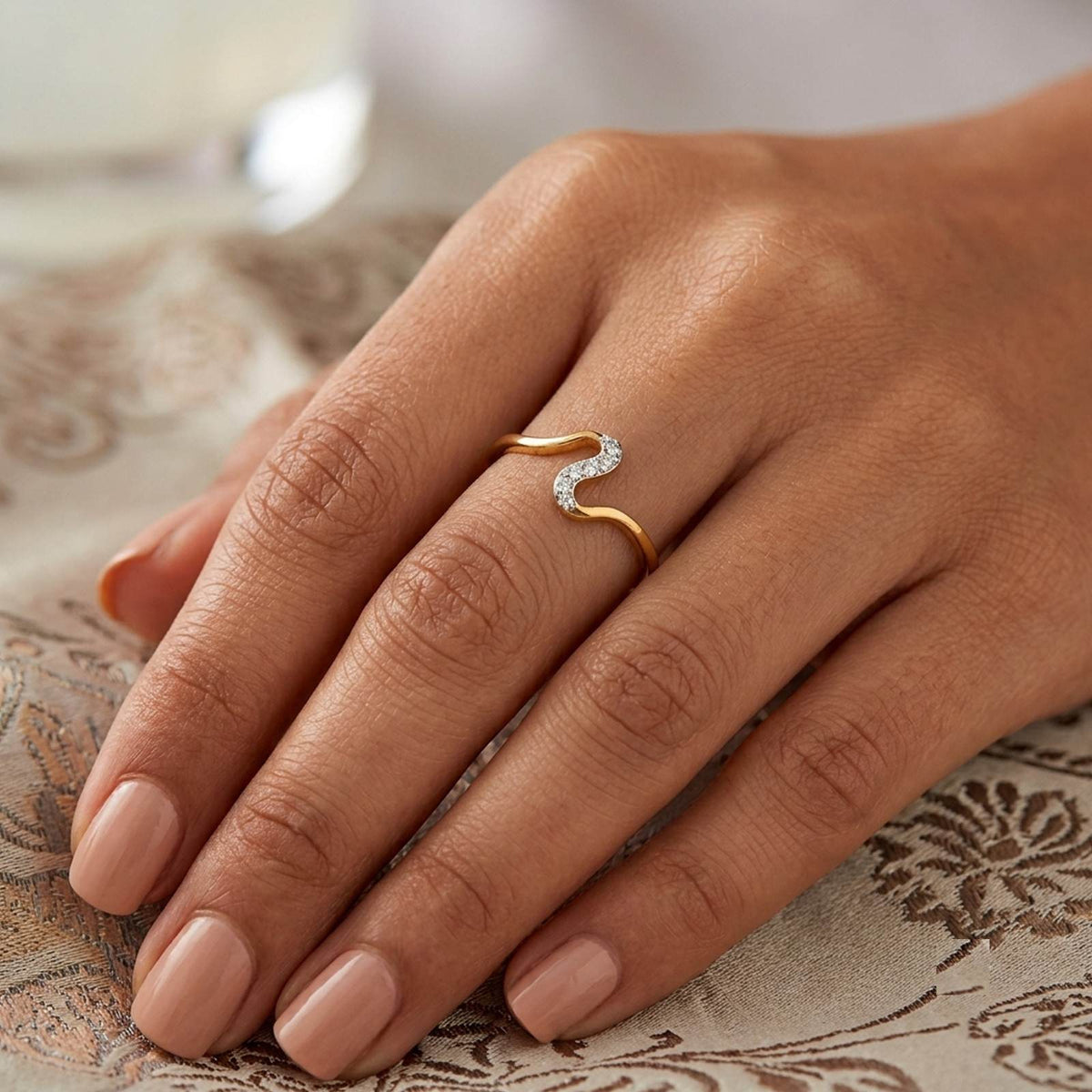 A close-up of a hand wearing a gold ring, resting on a patterned beige fabric surface.
