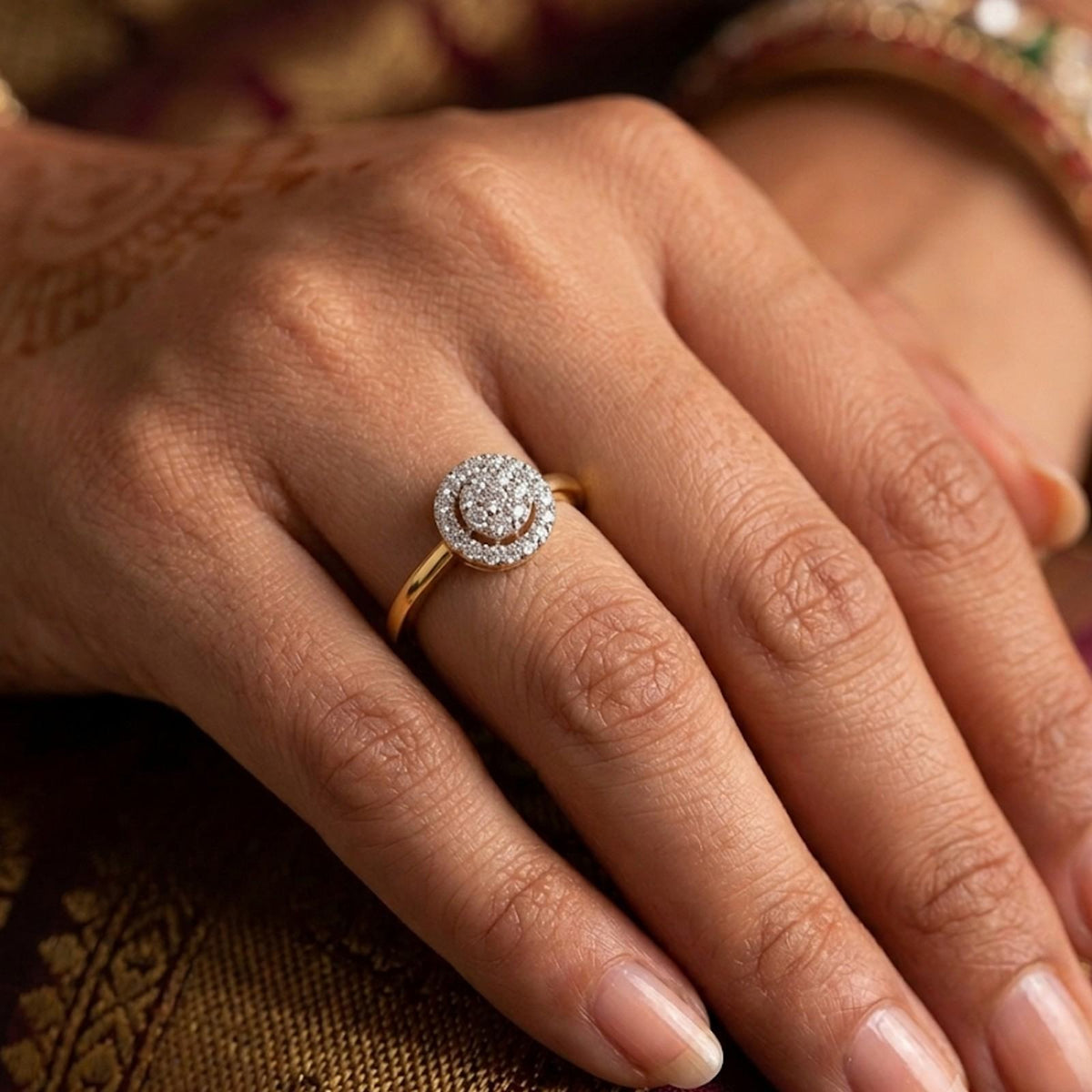 A close-up of a hand wearing a gold ring with a round diamond halo, resting on a patterned gold fabric surface.