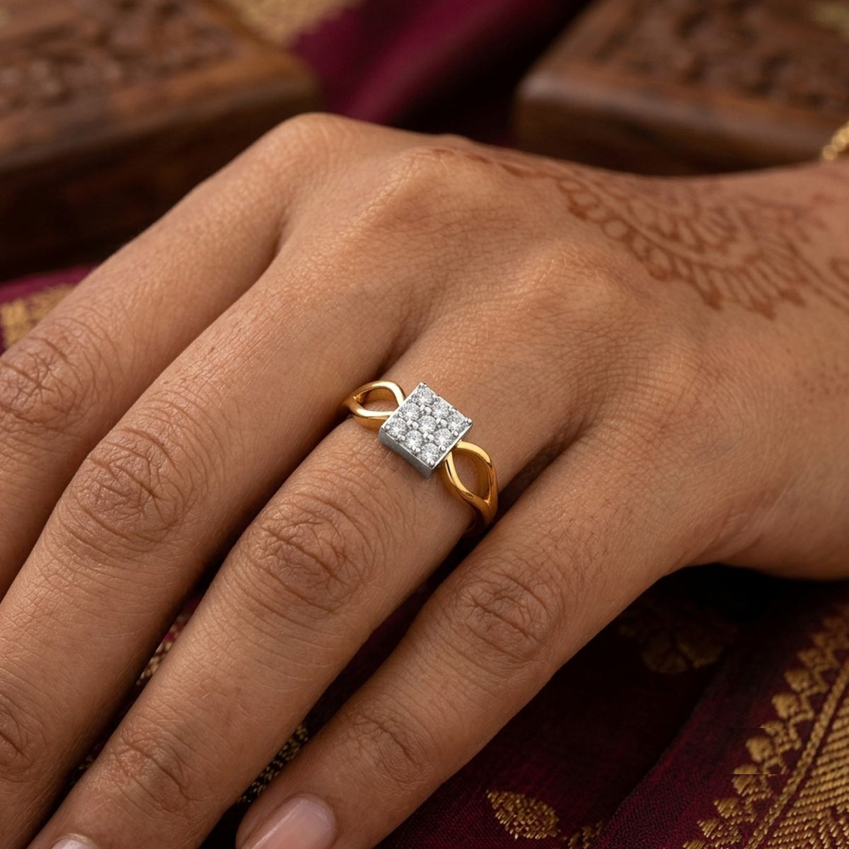 A gold ring with a square diamond cluster setting worn on a hand, set against a dark maroon saree.