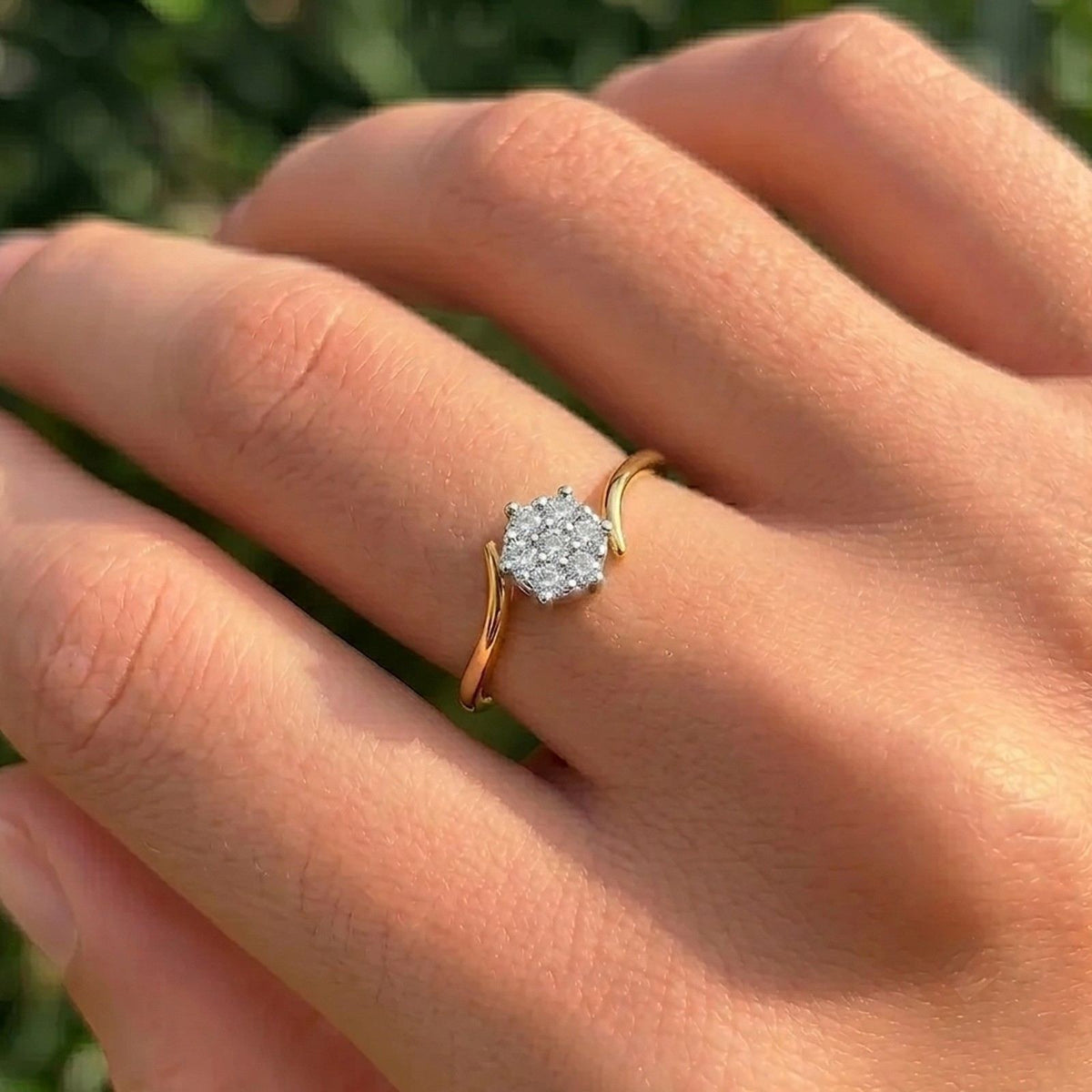A close-up of a hand wearing a yellow gold ring with a cluster of clear gemstones, set against a soft, blurred green background.