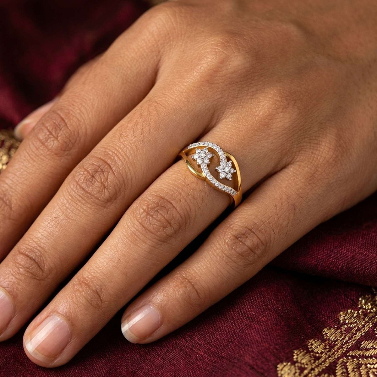A close-up of a hand wearing a gold ring with a flower motif set, resting against a deep maroon fabric background.