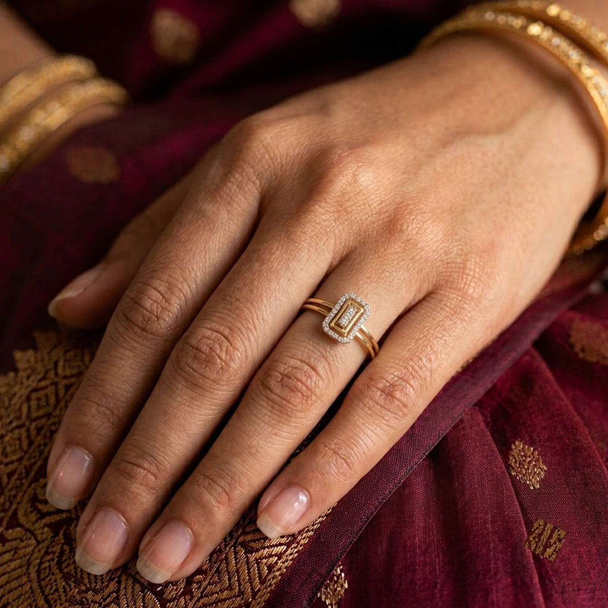 A close-up of a hand wearing a gold ring with a rectangular, resting on a deep maroon fabric background.