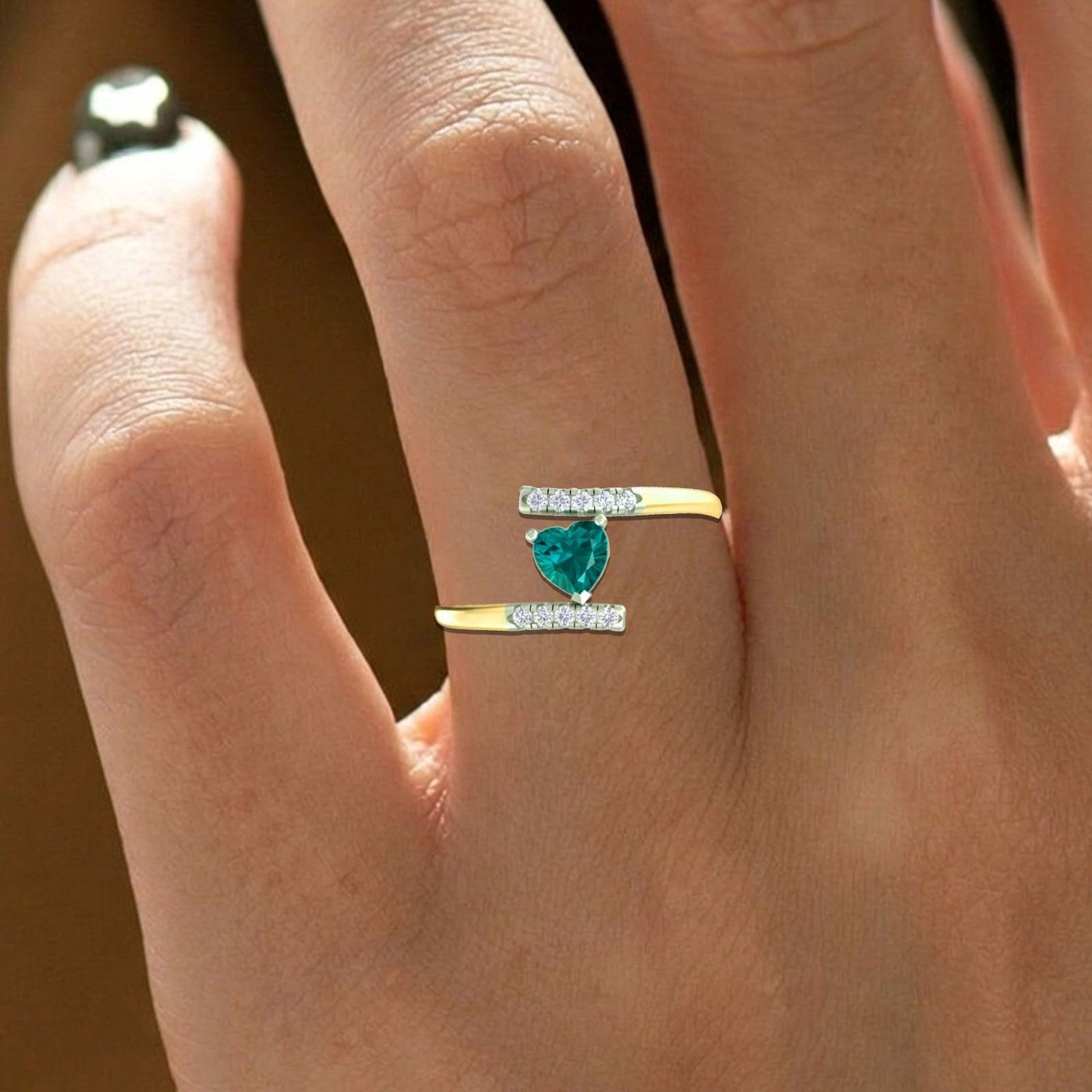 A close-up of a hand wearing a gold ring with a heart-shaped green gemstone on a warm brown background.