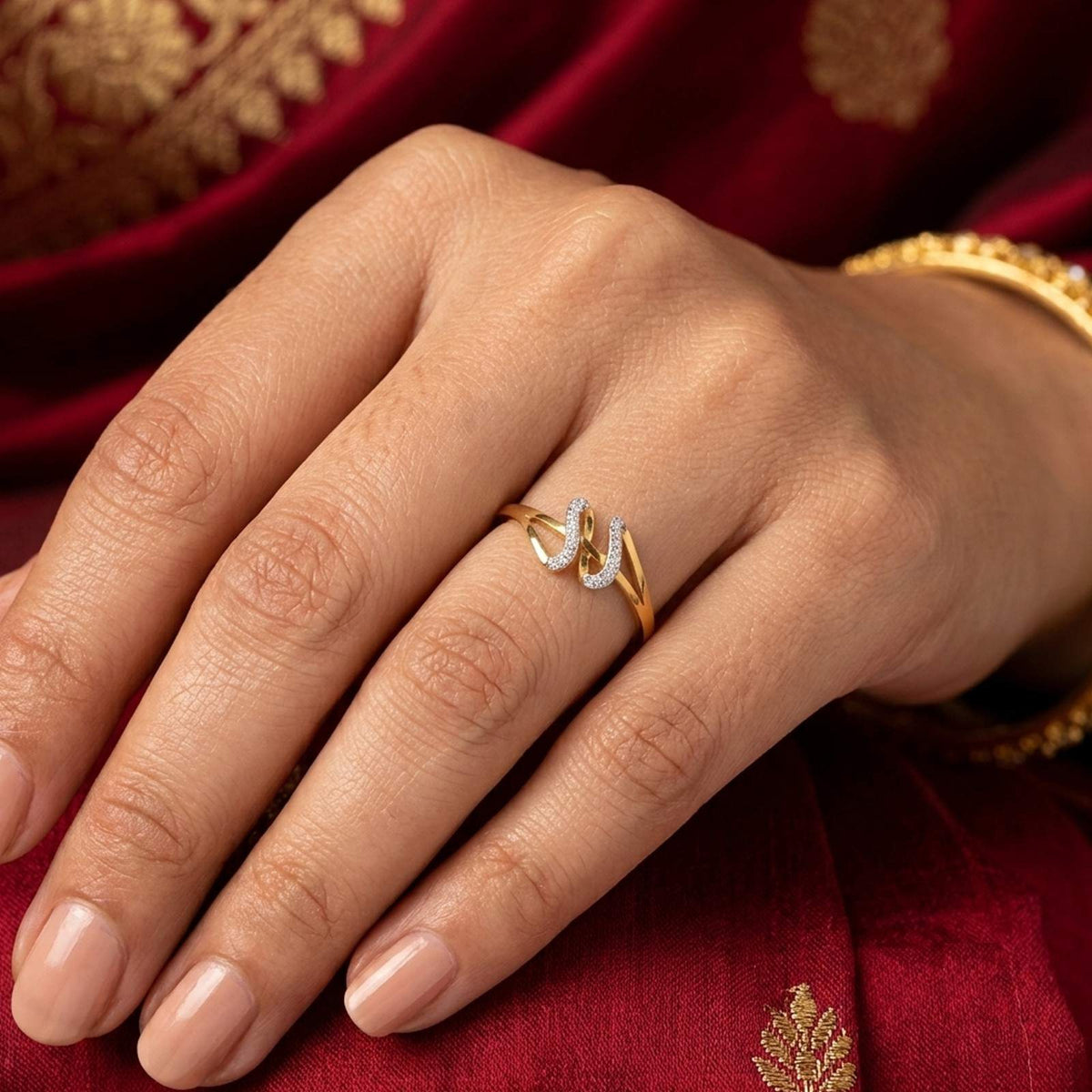 A close-up of a hand wearing a gold ring with a curved, resting on a deep red embroidered fabric background.