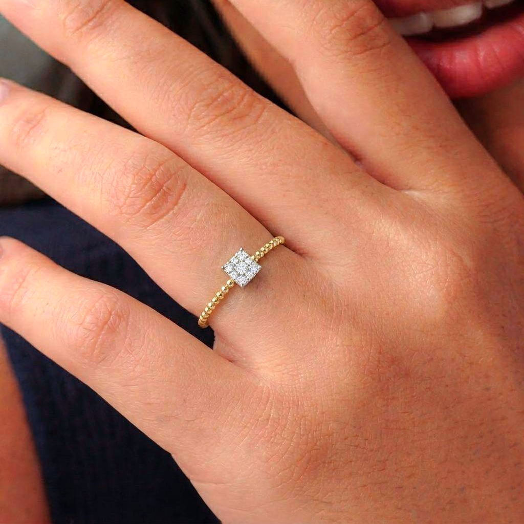 A close-up of a hand wearing a delicate gold ring with a small square cluster of clear stones on a textured gold band.