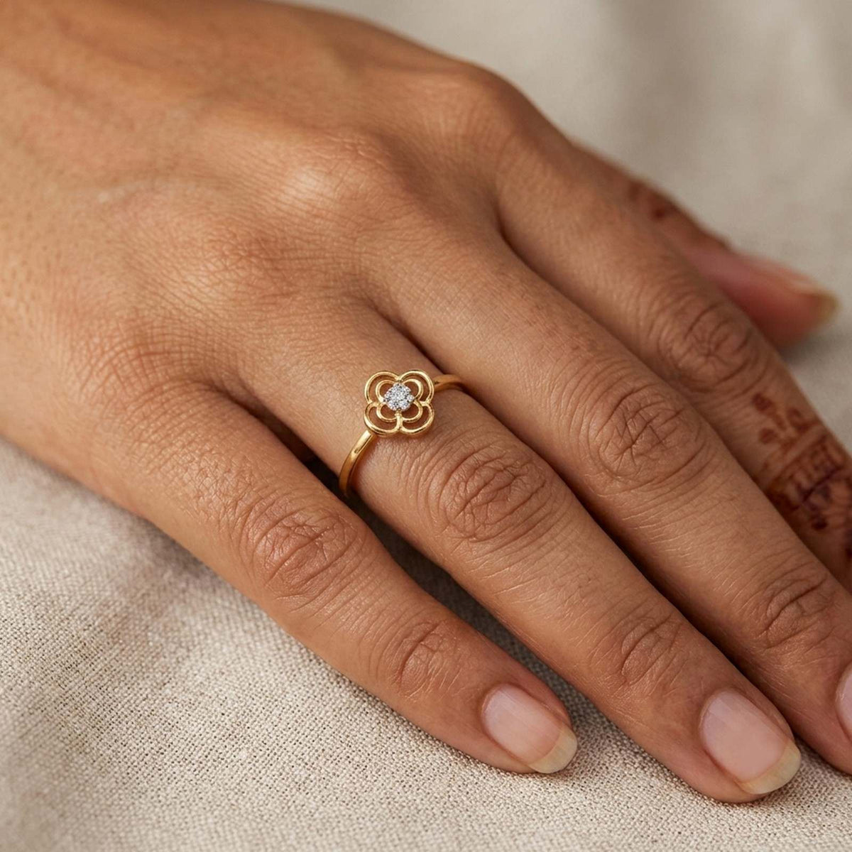 A gold ring with a small flower-shaped setting, worn on a person's finger against a soft beige fabric background.
