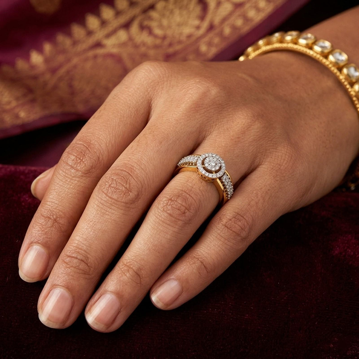 A close-up of a hand wearing a gold halo ring with a round diamond center, on a deep maroon velvet surface.