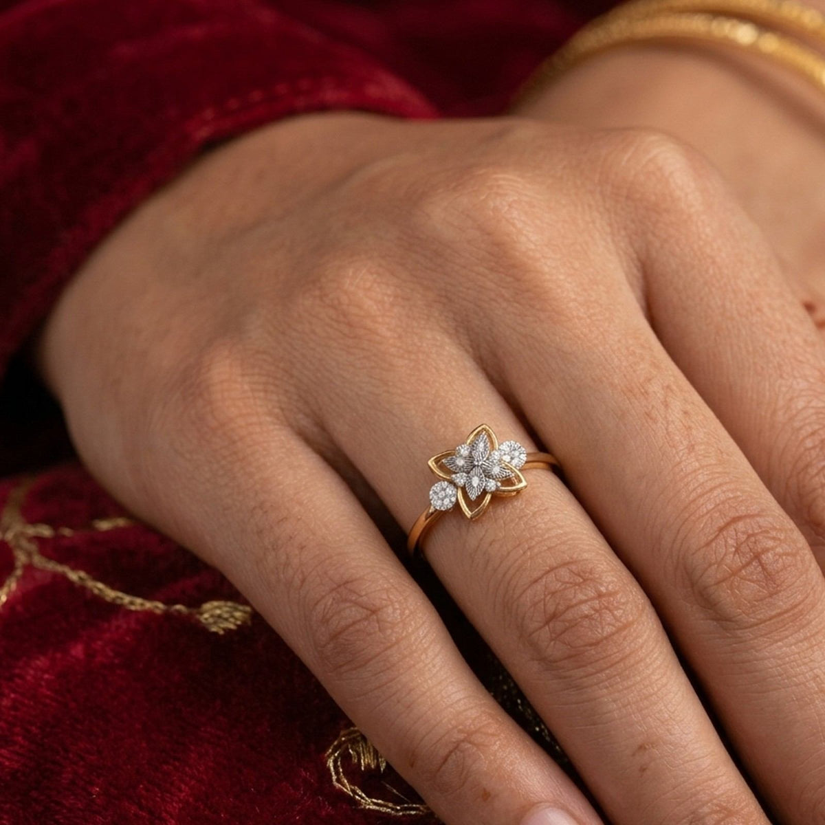 A gold ring with a flower-shaped cluster, worn on a person's finger. The hand is resting on a deep red velvet fabric.