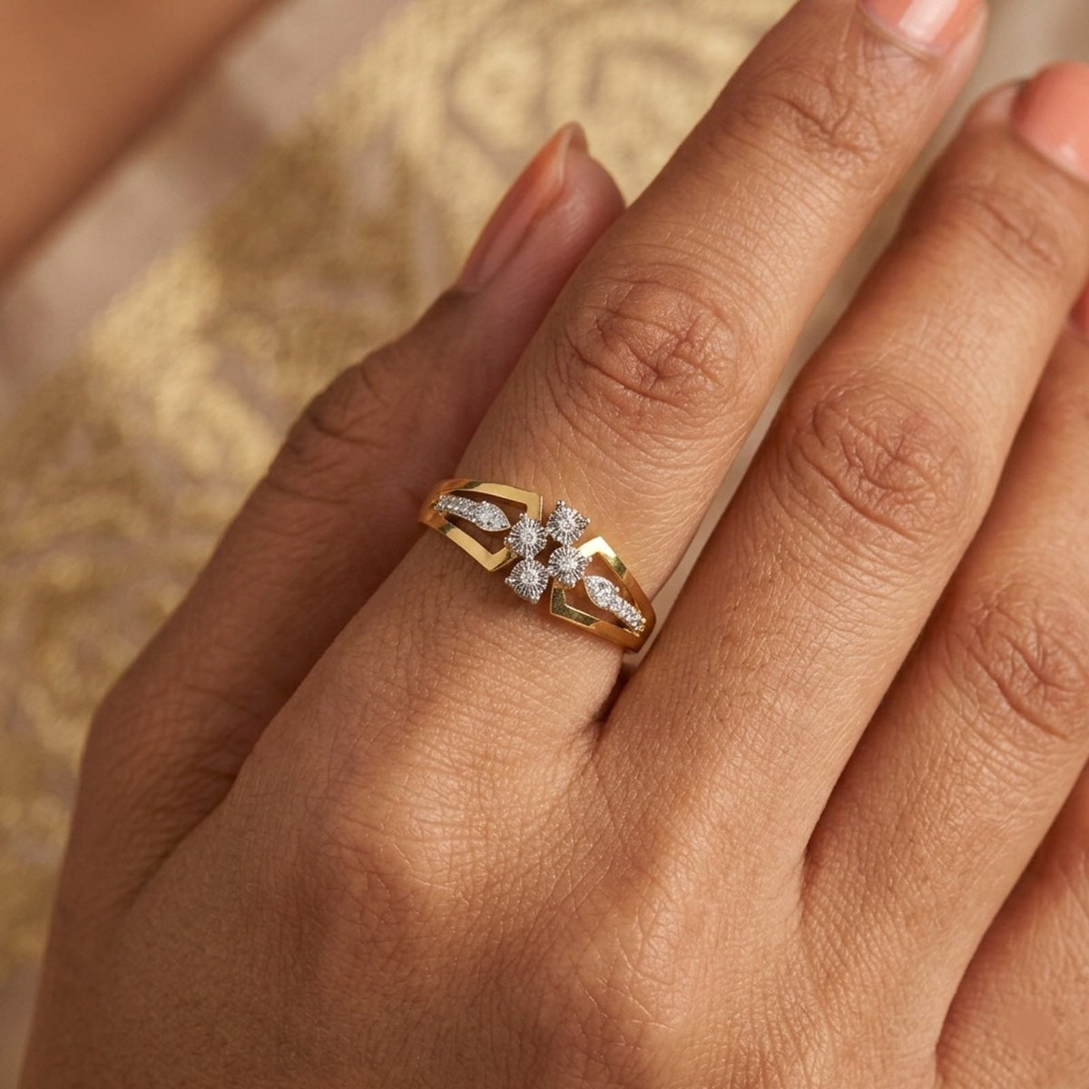 A close-up of a hand wearing a yellow gold ring with a diamond flower motif, set against a soft, warm patterned background.