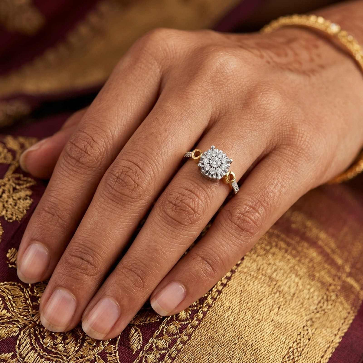 A close-up of a hand wearing a gold and diamond floral cluster ring, set against a rich gold and maroon patterned fabric background.