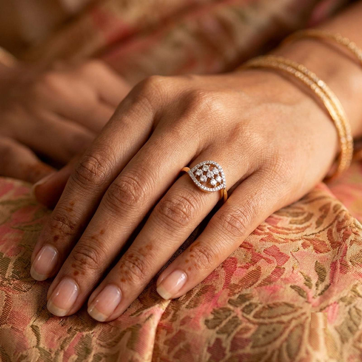 A close-up of a hand wearing a gold ring with a diamond-patterned oval face, resting on a pink and gold patterned fabric surface.