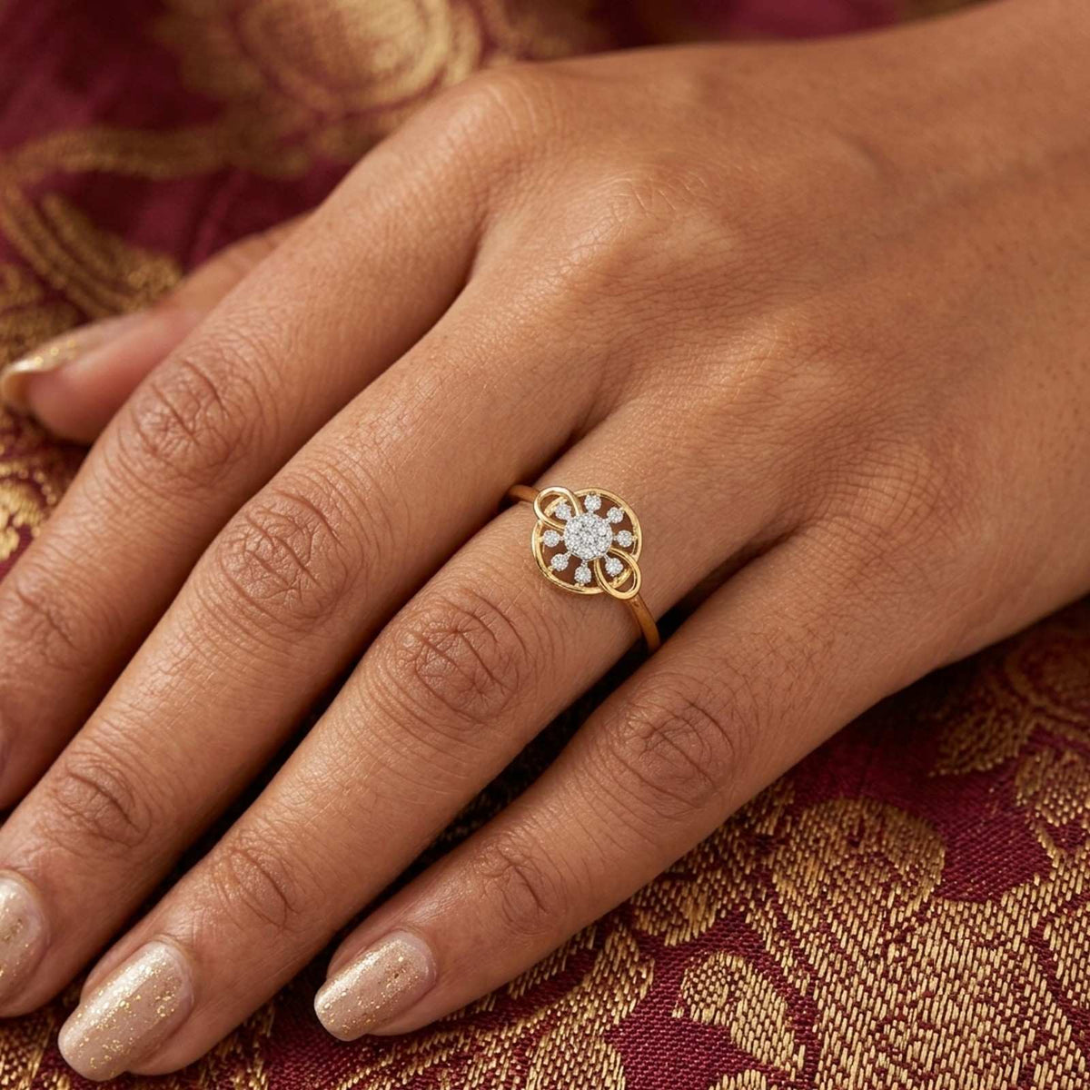 A close-up of a hand wearing a gold ring with a floral cluster setting, set against a rich burgundy and gold patterned fabric.