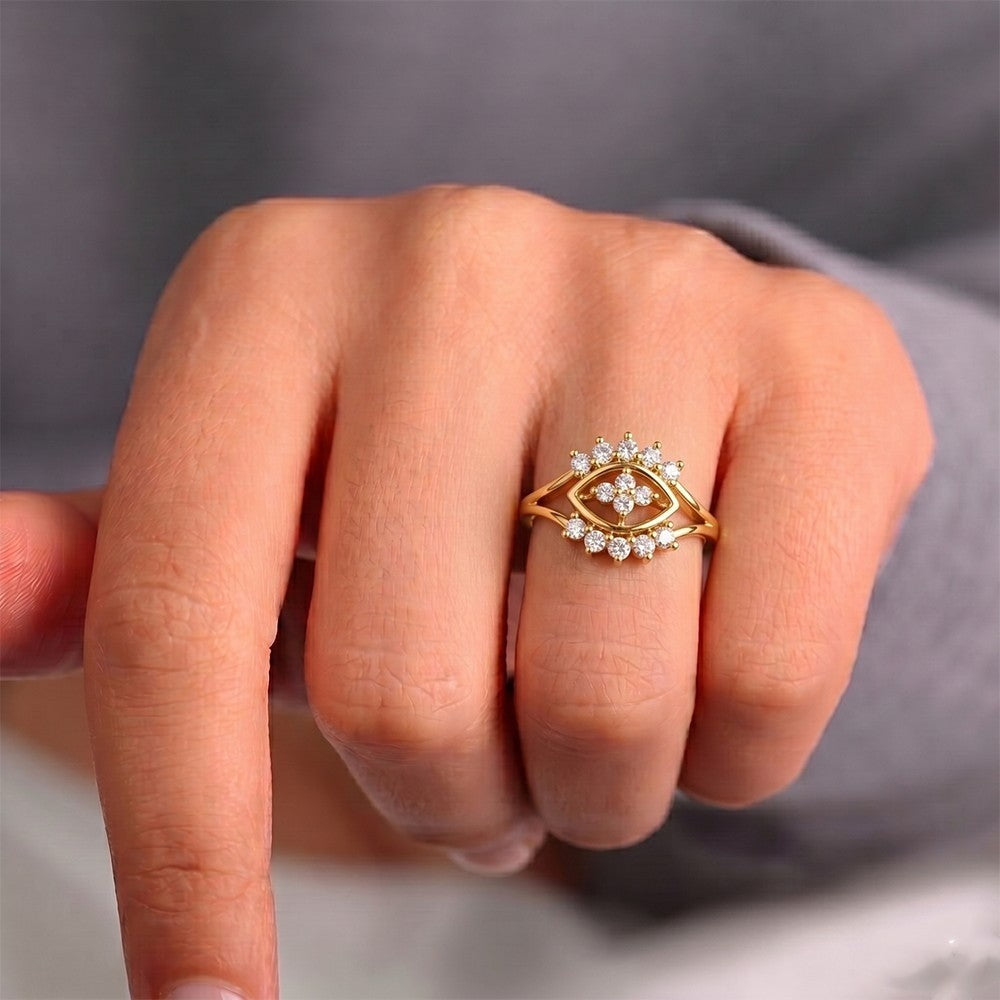A gold ring with a central eye motif, worn on a person's finger against a soft gray background.