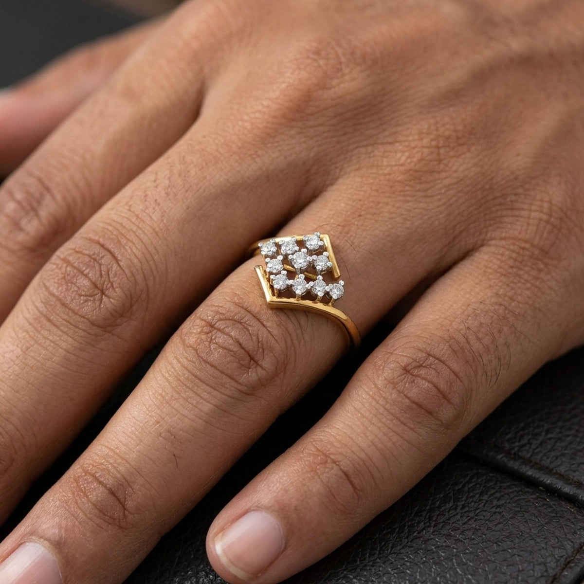 A close-up of a hand wearing a gold ring with a square cluster, on the ring finger, set against a dark textured background.