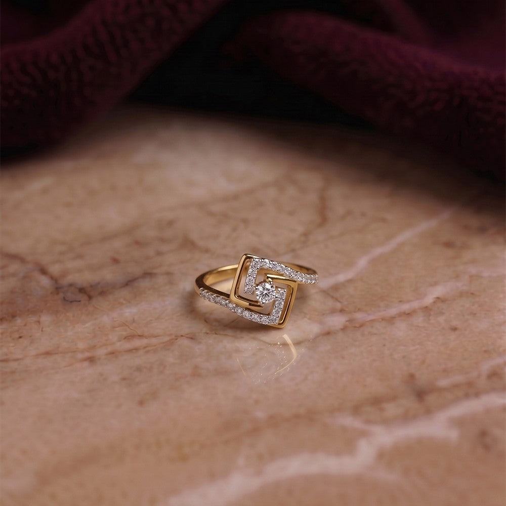 A gold and diamond geometric ring with pavé-set clear stones, resting on a beige marble surface with a dark burgundy fabric background.
