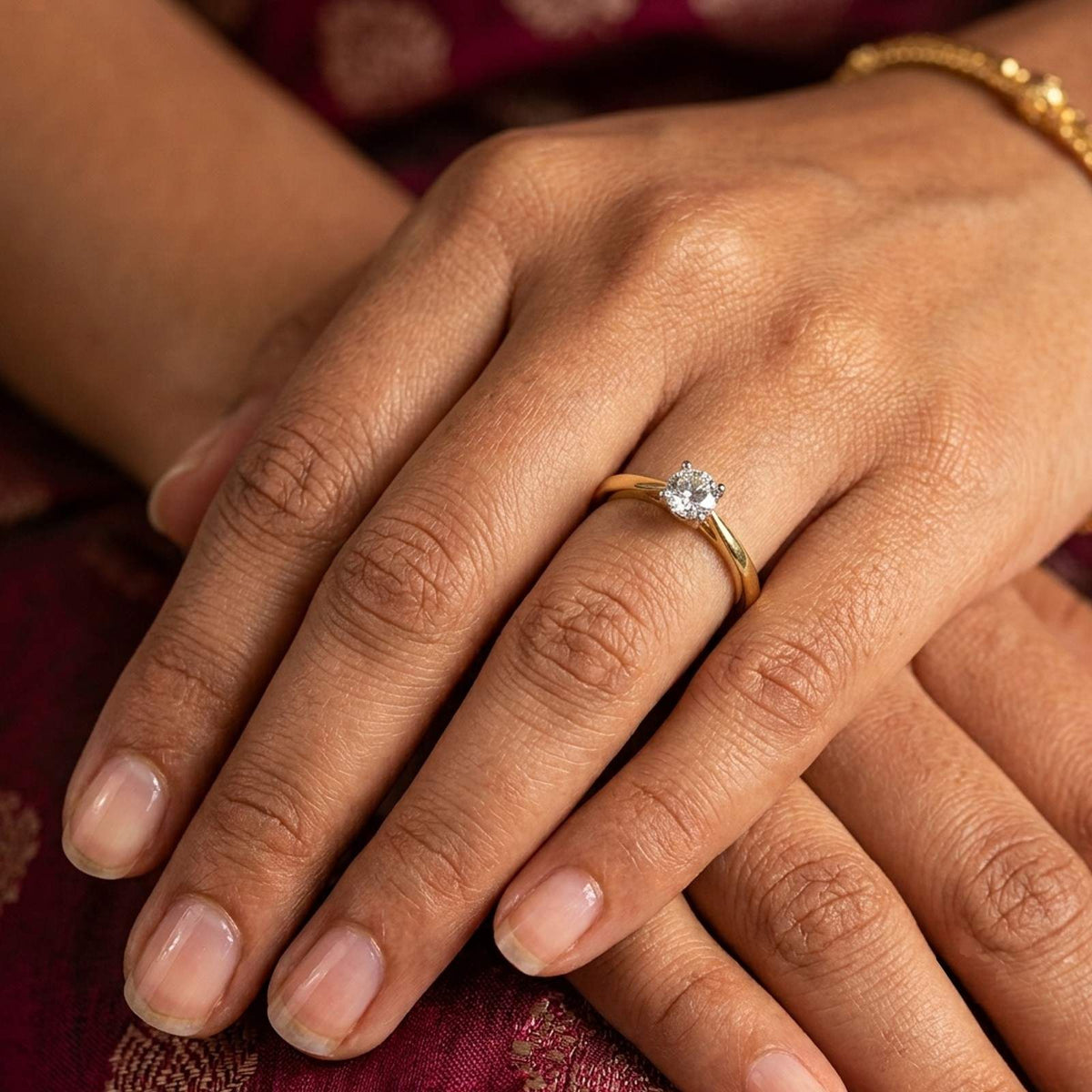 A close-up of a person's hand wearing a gold solitaire ring, resting on a dark red patterned fabric.