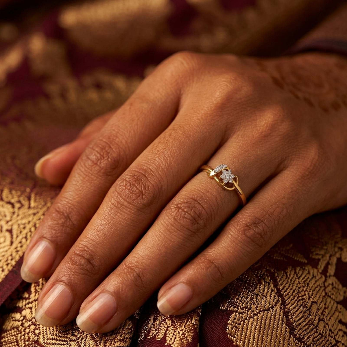 A close-up of a hand wearing a delicate gold ring, resting on a richly textured gold and burgundy fabric.