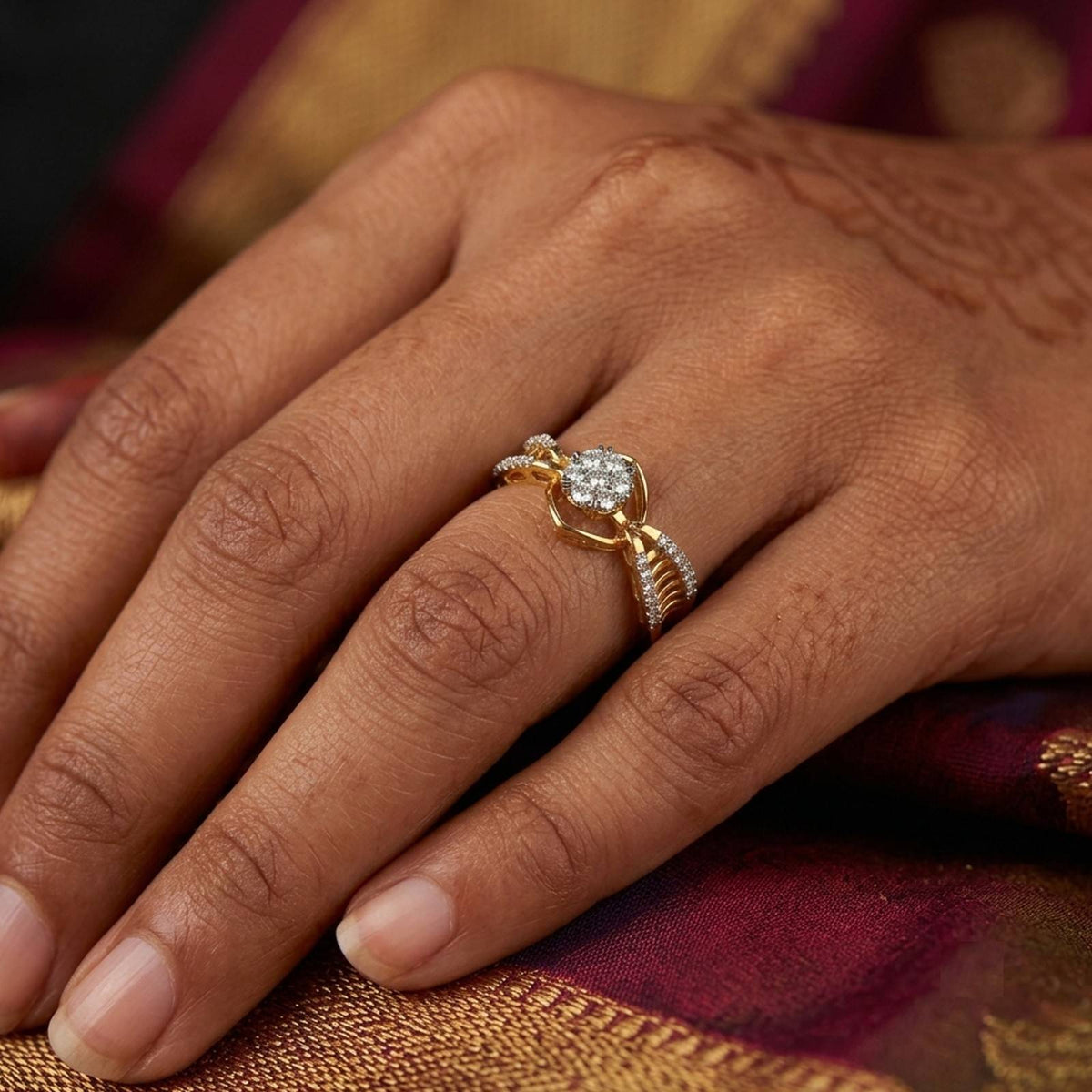 A close-up of a hand wearing a gold ring with a round cluster center, resting on a rich burgundy and gold fabric surface.