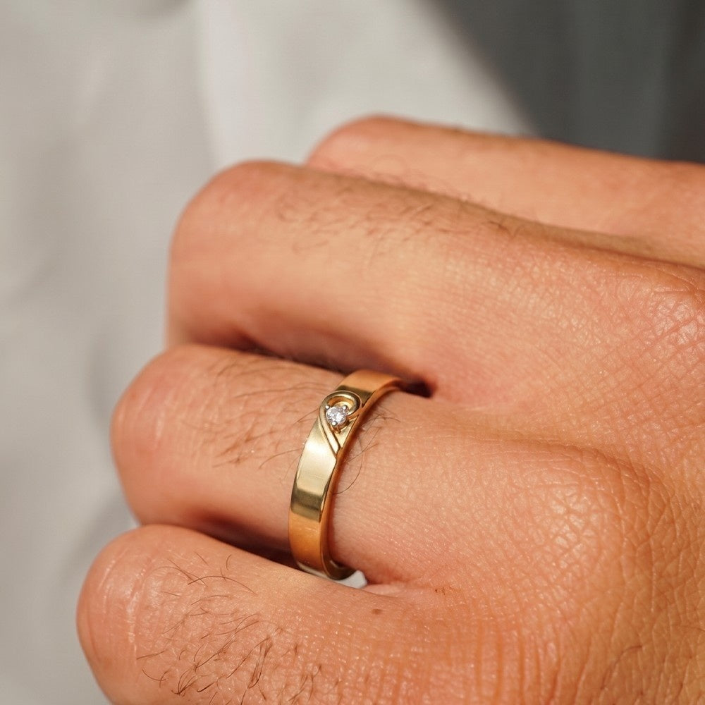 A close-up of a hand wearing a gold band ring with a small round clear diamond set in a curved setting.