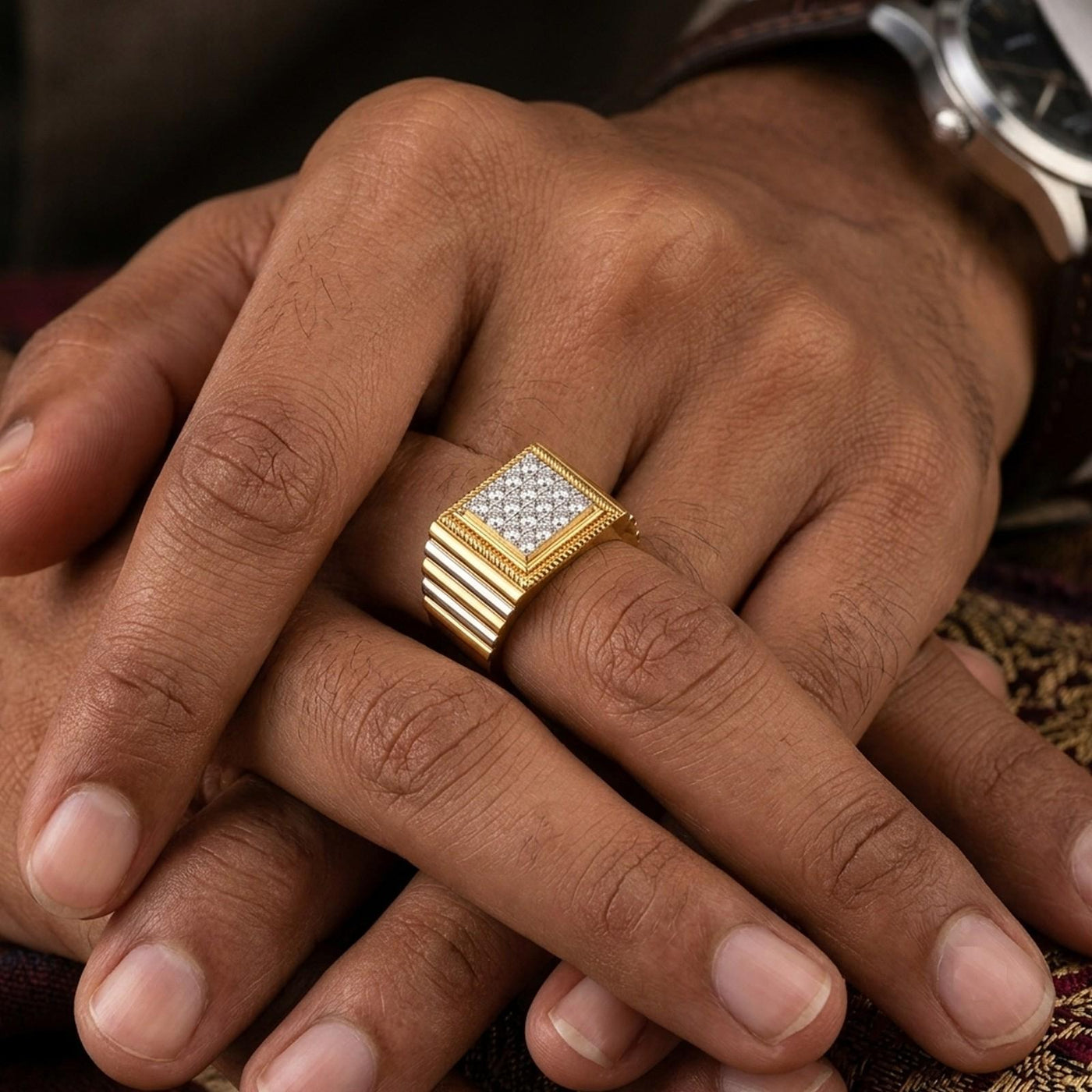 A close-up of a man's hands resting on each other, wearing a gold square ring with a diamond-encrusted face on one finger.
