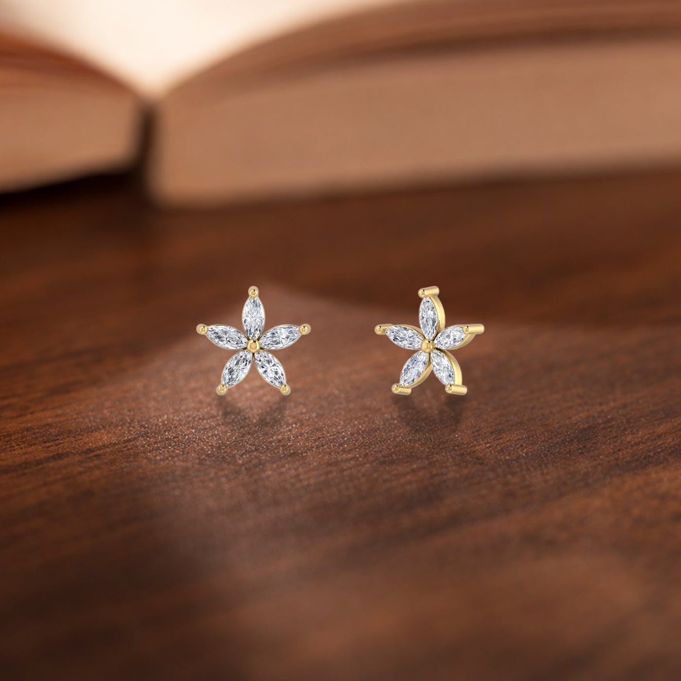 A pair of gold flower-shaped stud earrings with marquise-cut clear stones, displayed on a dark wooden surface with a blurred open book in the background.