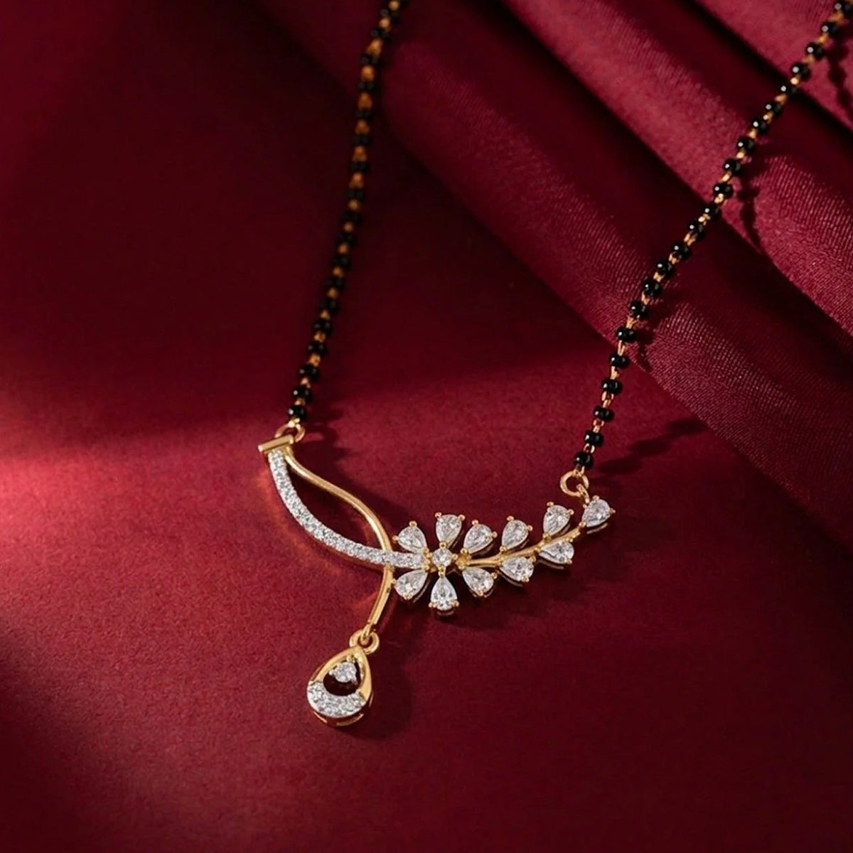 A gold chain mangalsutra with black beads and an asymmetrical diamond featuring floral and curved motifs, resting on dark red fabric