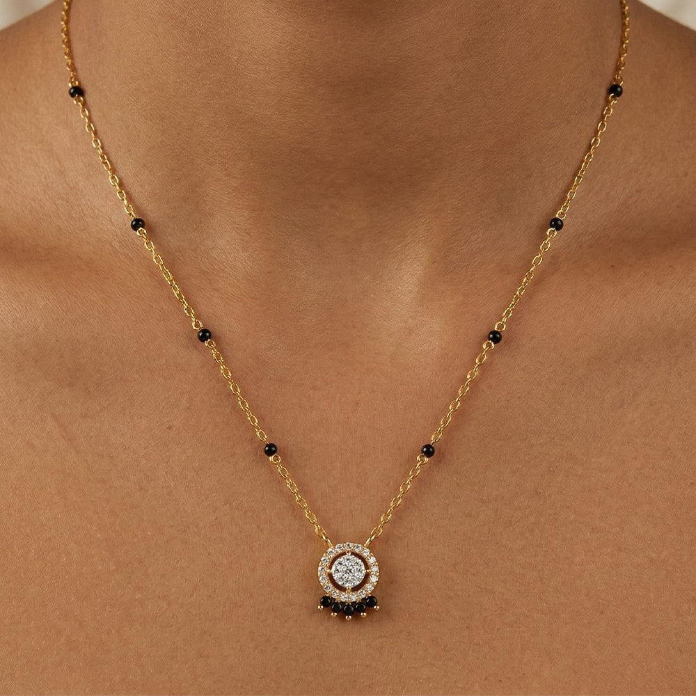 A gold and diamond mangalsutra with small black bead accents and a circular featuring a clear center stone and black surrounding stones, shown worn on a model's neck.