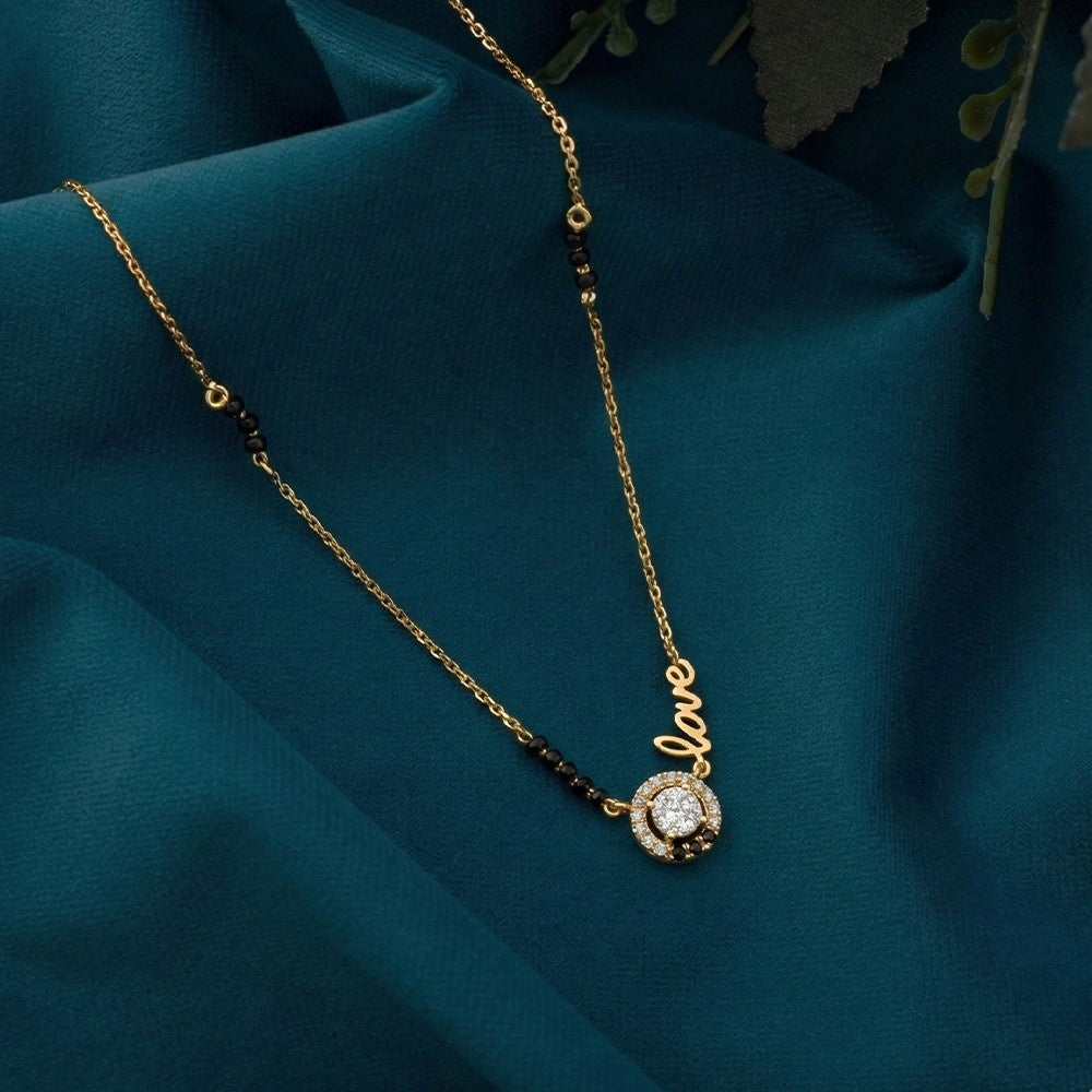 A yellow gold and diamond mangalsutra with a script 'love' pendant, a small round clear stone, and dark bead accents, displayed on draped teal fabric.