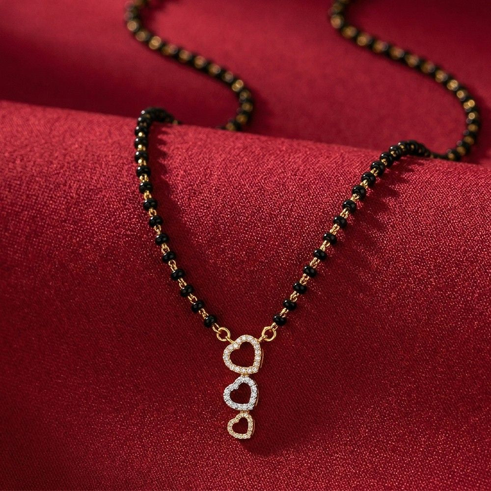 A gold and diamond mangalsutrawith black and gold beads and a gold featuring three stacked heart-shaped crystal clusters, displayed on a textured red fabric background.